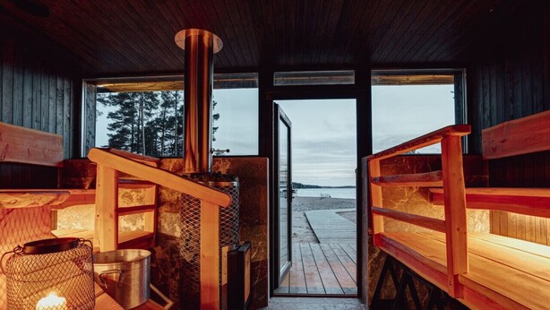 A wooden interior with a large window leading to a beach view, a metal stove, and a lantern. - Saunamäki Resort Guesthouse - Countryside Views (Salo)