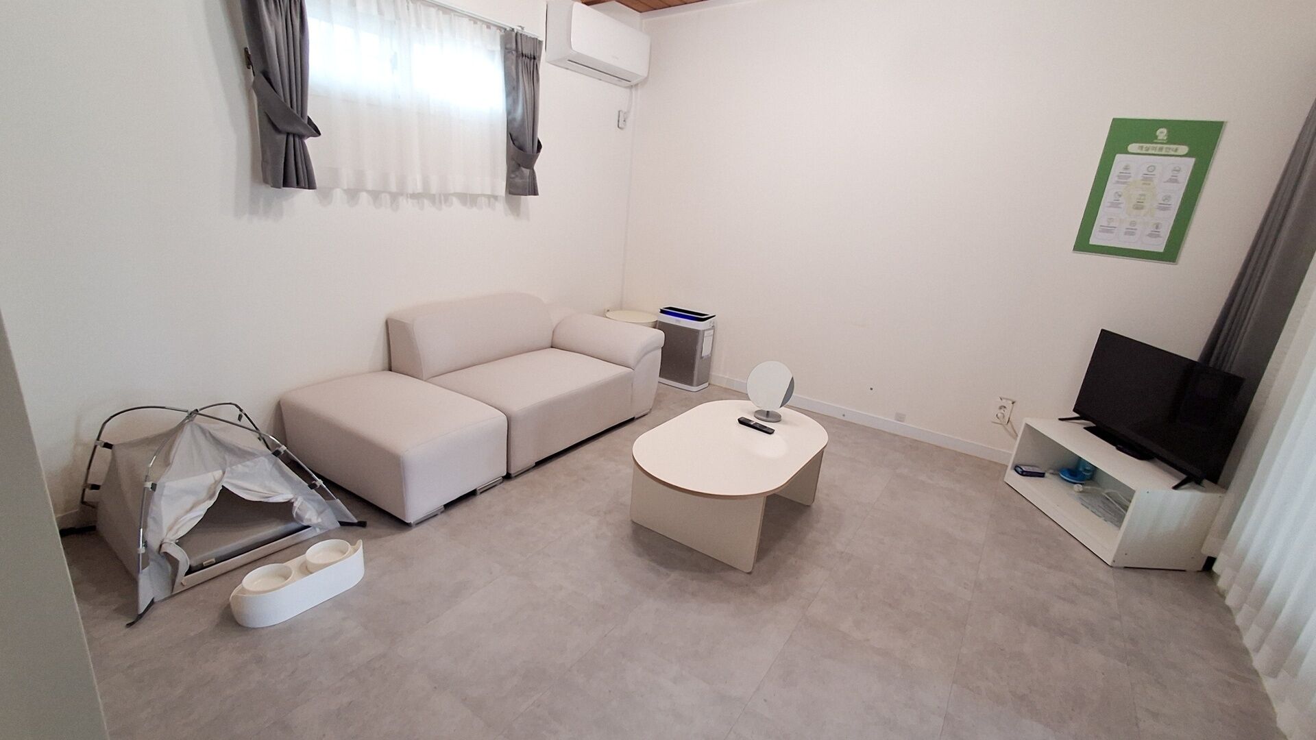 A small, well-lit room with a sofa, a small table, a TV, and a window with curtains.