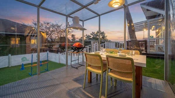 Eine verglaste Terrasse mit Tisch, Stühlen, einem Grill und Blick auf die Umgebung.