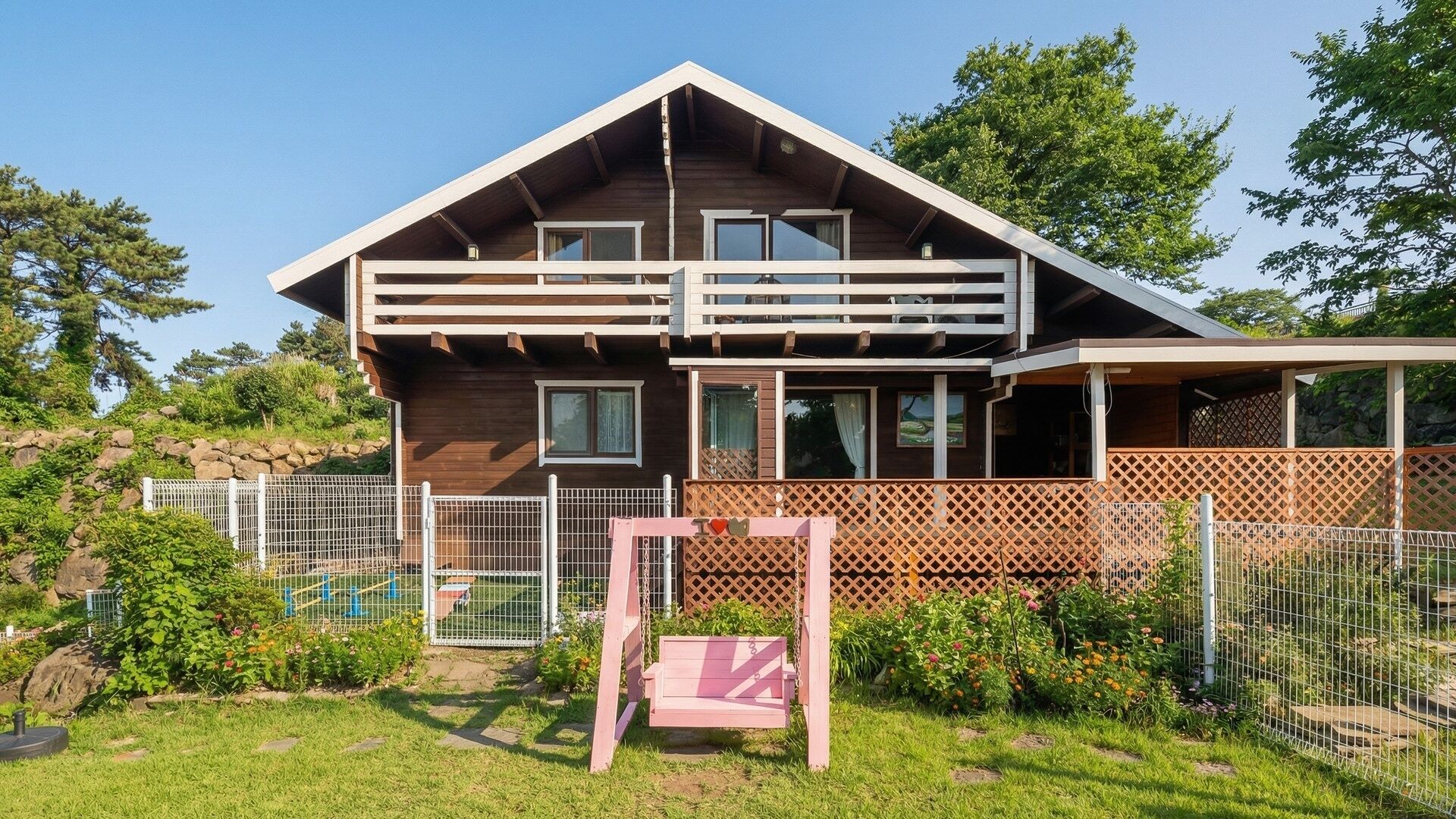 A two-story wooden house with a balcony, surrounded by a fenced garden with various plants and a swing.
