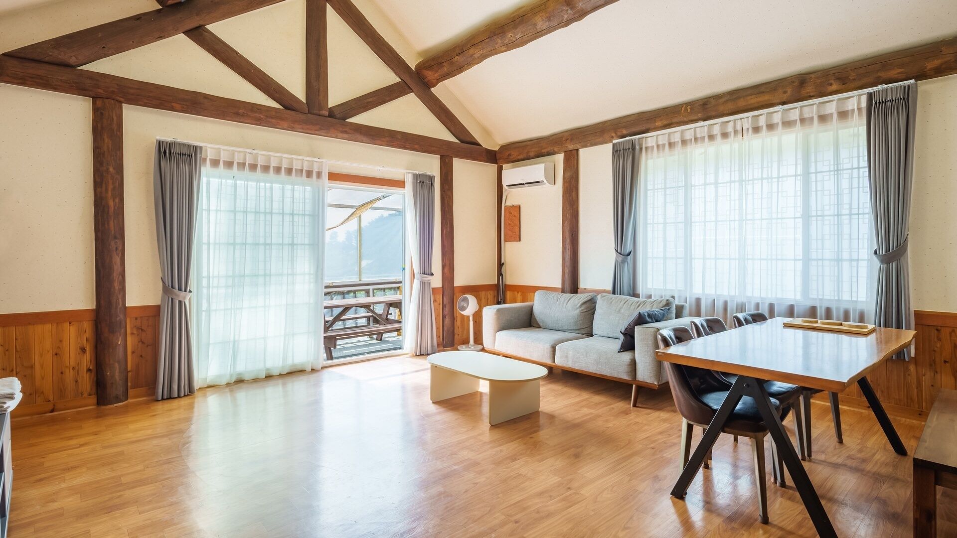 A spacious living area with a wooden floor, a sofa, a dining table, and a balcony with a view.