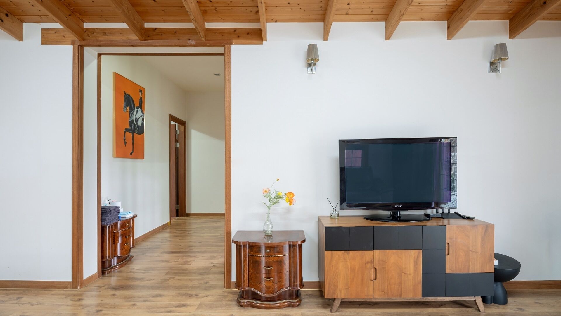 Een moderne woonkamer met een televisie op een houten kast, een klein bijzettafeltje met een vaas, en een schilderij aan de muur.