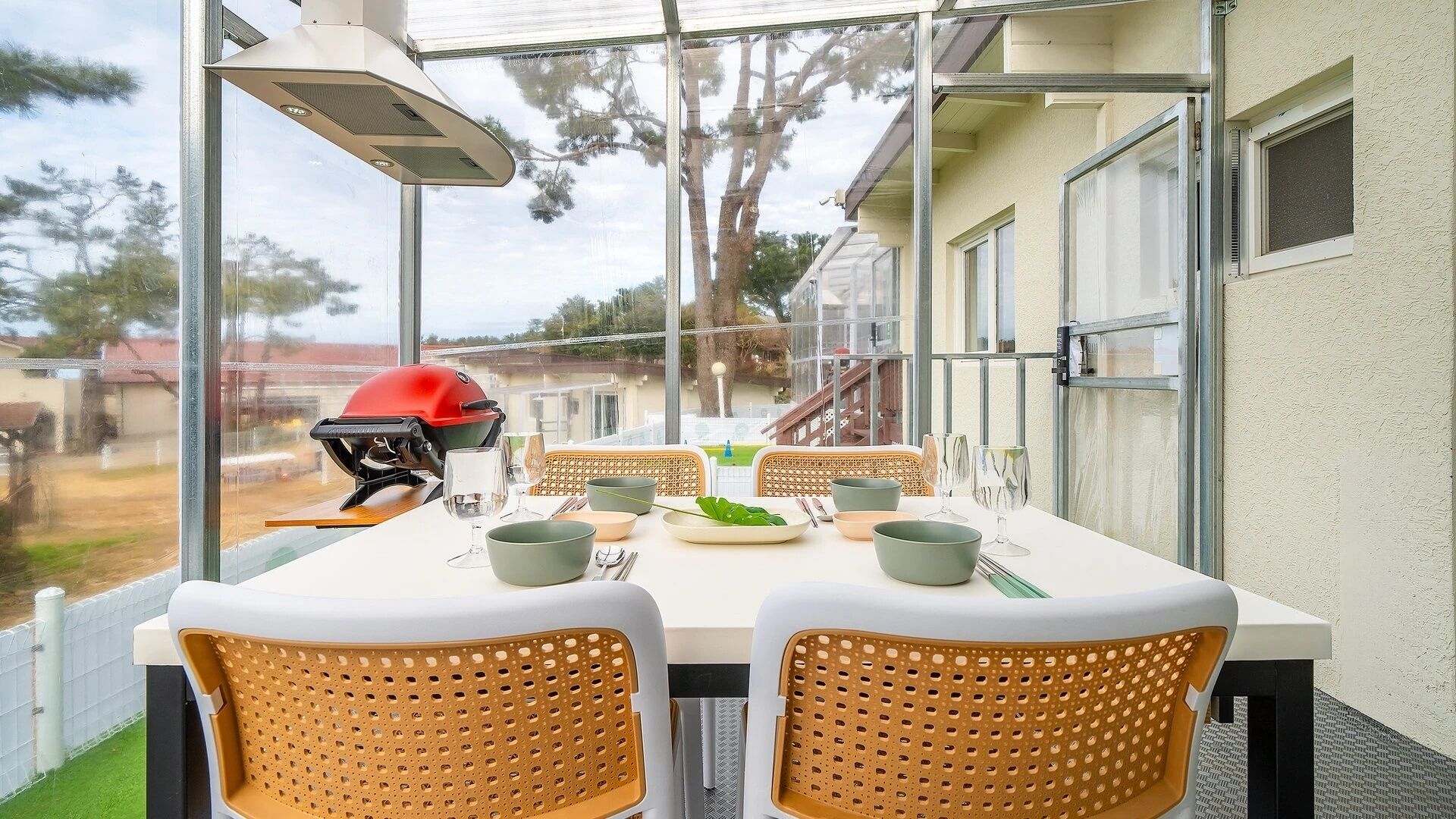 Un espace extérieur aménagé pour les repas, avec une table dressée pour deux personnes, entouré de chaises en osier, d’un barbecue et offrant une vue sur un quartier résidentiel.