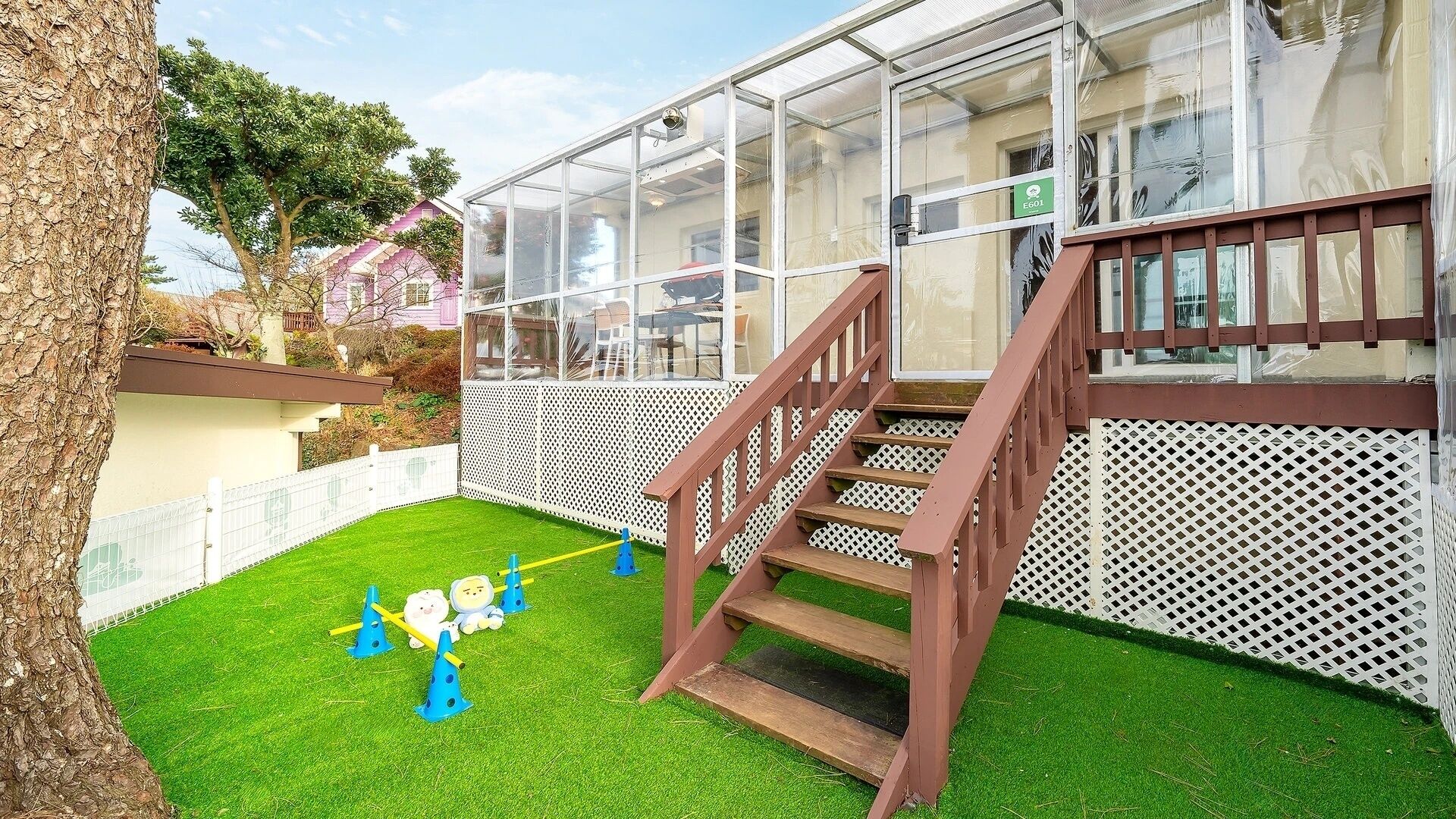 A backyard with artificial grass, a wooden staircase, a play area with a slide, and a glass-enclosed structure.