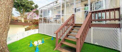 A backyard with artificial grass, a wooden staircase, a play area with a slide, and a glass-enclosed structure.
