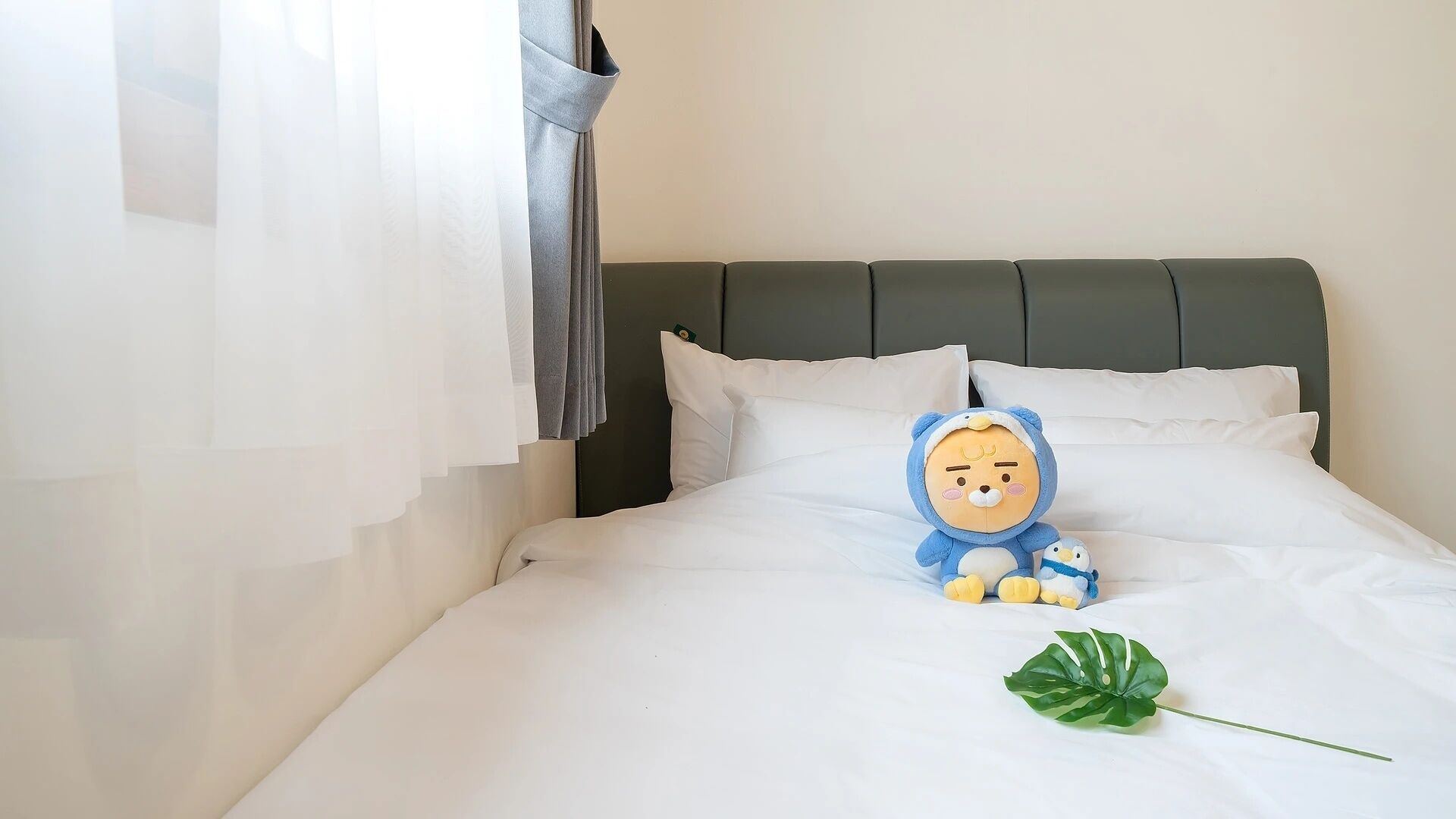 Una cama con ropa de cama blanca, un peluche y una planta verde.