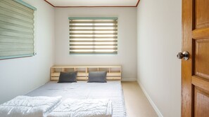 A bedroom with a bed, two pillows, a window with blinds, and a wooden door. - Jeju Dangdangi Park Resort Pension (Jeju City)
