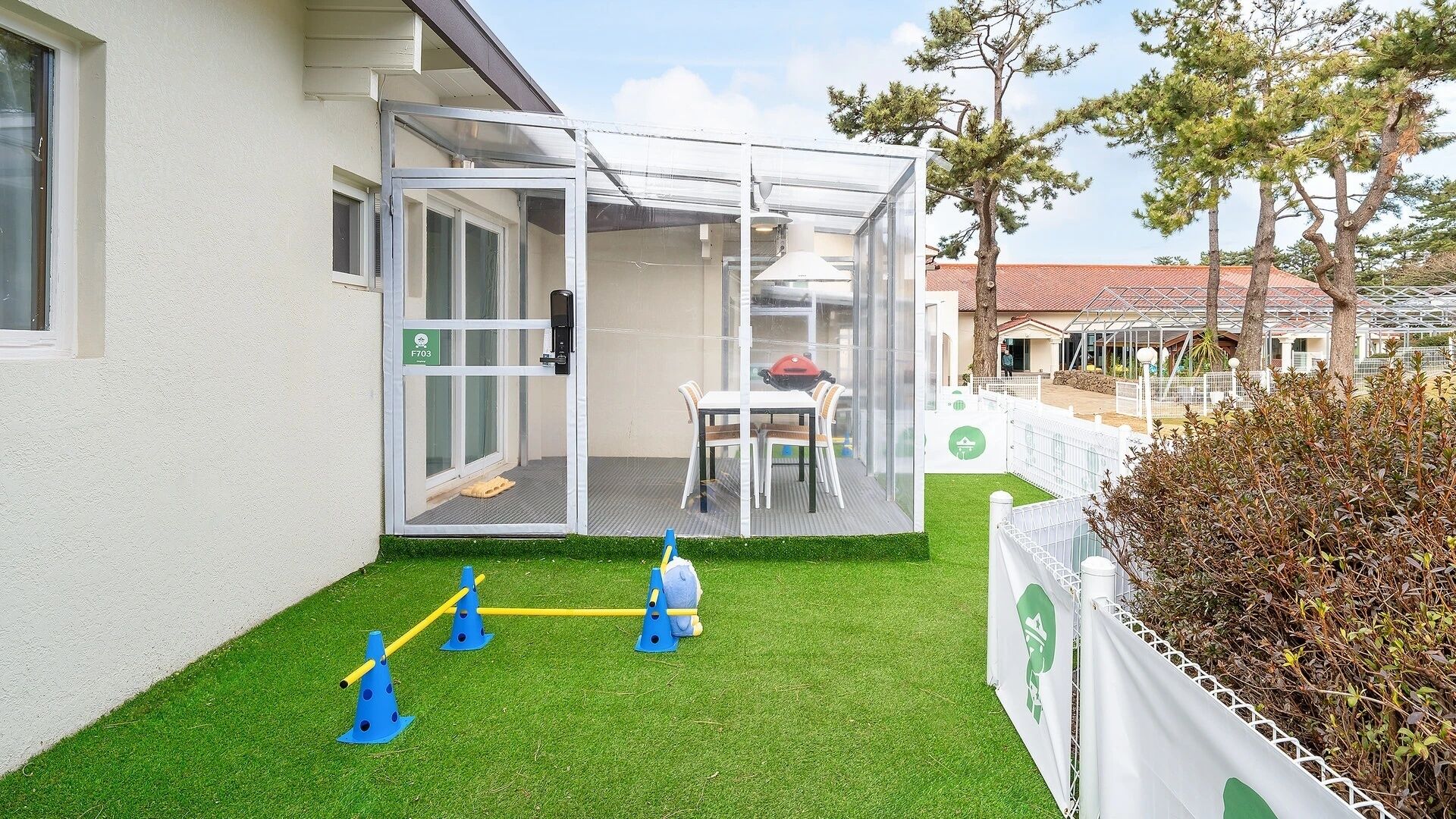 Un espacio al aire libre techado con puerta de vidrio, una mesa y sillas, y un área de juegos pequeña con conos y pelota.