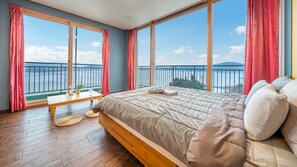 Un dormitorio con una cama grande, una mesita de noche de madera y vistas al mar a través de amplios ventanales.