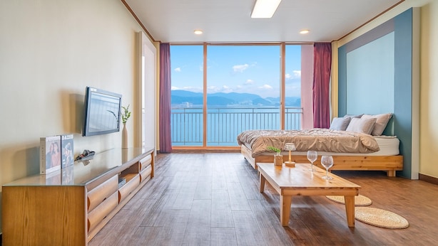 A modern hotel room with a large bed, a TV, a wooden desk, and a balcony with a view of the sea. - Daebudo Green Beach Pension (Ansan)