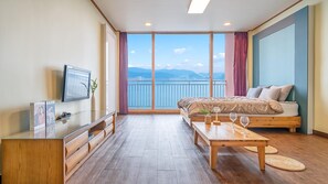 A modern hotel room with a large bed, a TV, a wooden desk, and a balcony with a view of the sea. - Daebudo Green Beach Pension (Ansan)