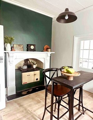 Uma sala de jantar com mesa de madeira escura, duas cadeiras, luminária pendente, lareira e parede verde, além de uma janela com persianas.