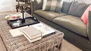 A wicker coffee table with books and a wine bottle, a grey sofa with cushions, and a window with natural light. - Telegraph Station Stays

 (Penola)