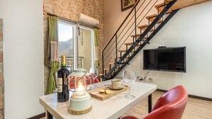 Un comedor con una mesa preparada para dos personas, una botella de vino, una vela y una bandeja con galletas. Se ve una escalera y un televisor al fondo.