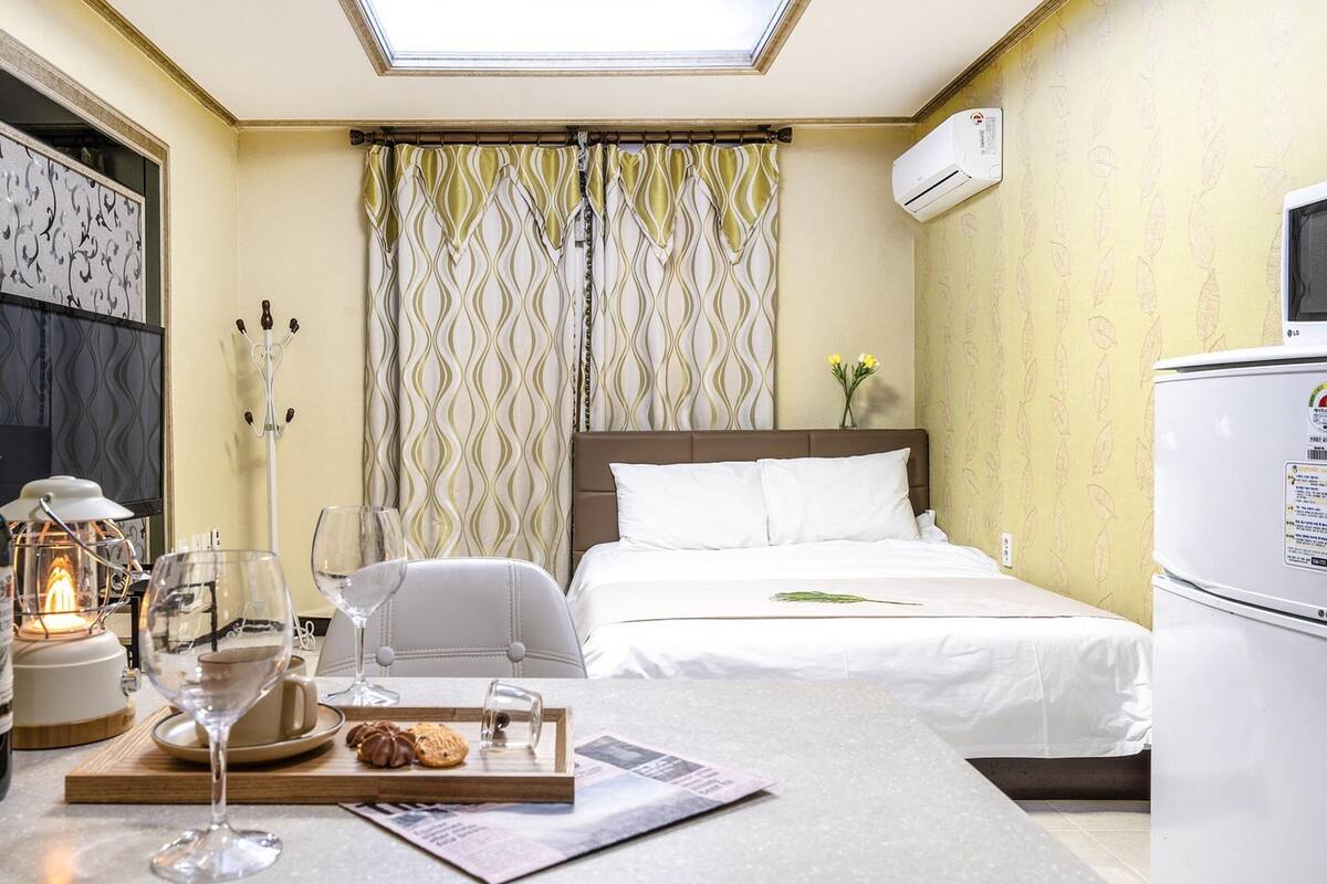 Ein Hotelzimmer mit einem Bett, einem Esstisch mit Wein, Gläsern und Gebäck, einem Sessel, einer Lampe und einer Klimaanlage.