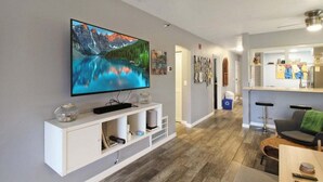 A modern living room with a flat-screen TV mounted on the wall, a white entertainment unit below, and a dining area with a table and chairs.