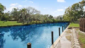 A swimming pool with a wooden deck and stone edging, surrounded by trees and a grassy area.