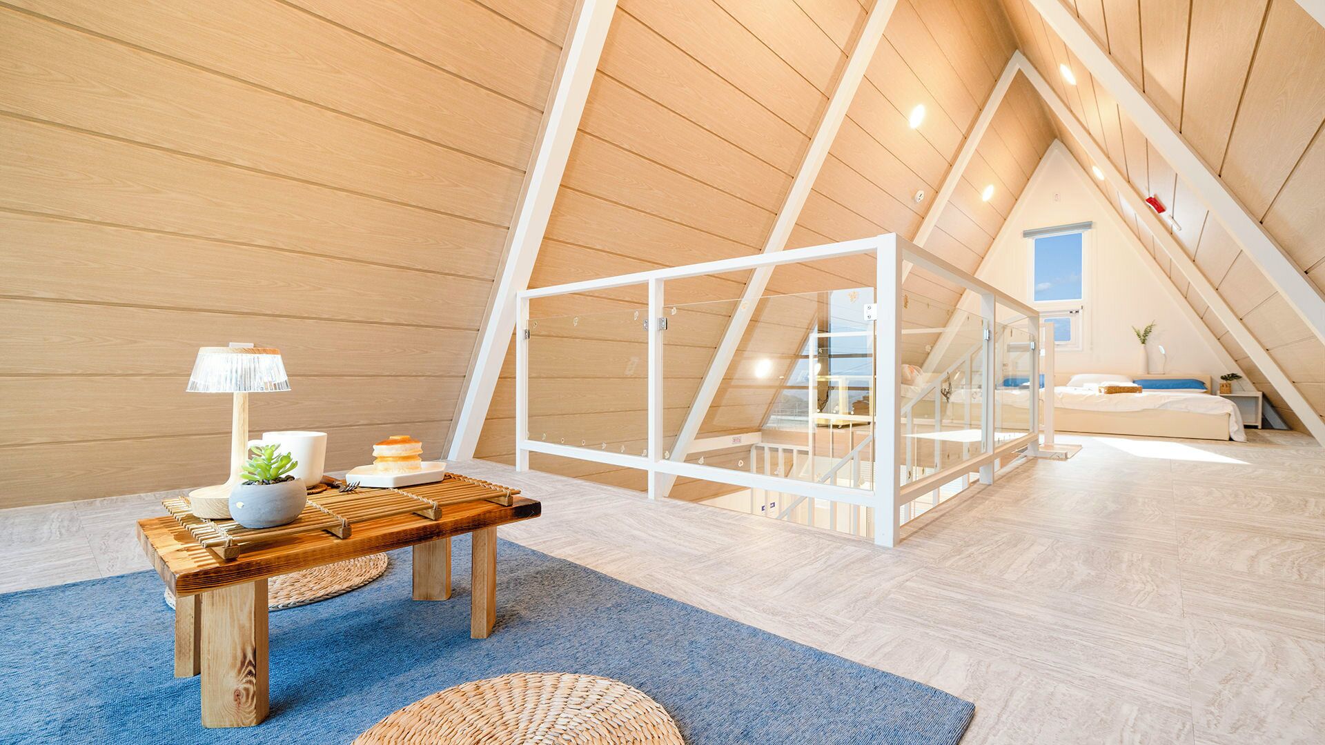 A modern loft bedroom with a sloped ceiling, a wooden table with a lamp, and a bed with a view of the outdoors.