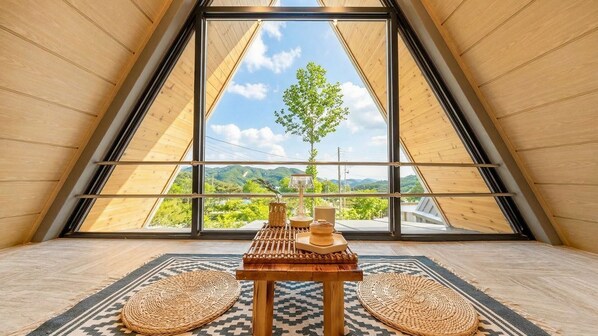 A room with a sloped ceiling, a large window with a view of greenery, a small table with a lamp, and woven mats on the floor. - Gapyeong The Paran Private Pool Villa (Gapyeong)