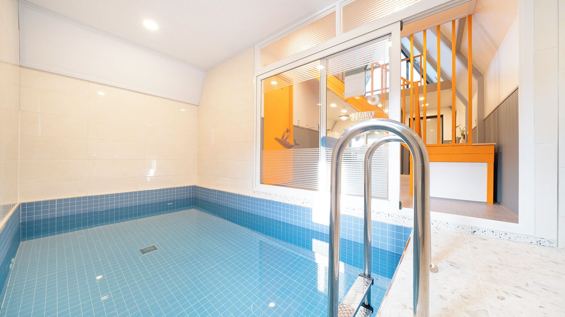 Ein Innenpool mit blau gefliestem Boden, einer Metallleiter und Blick auf eine Treppe, die zu einer oberen Ebene führt.