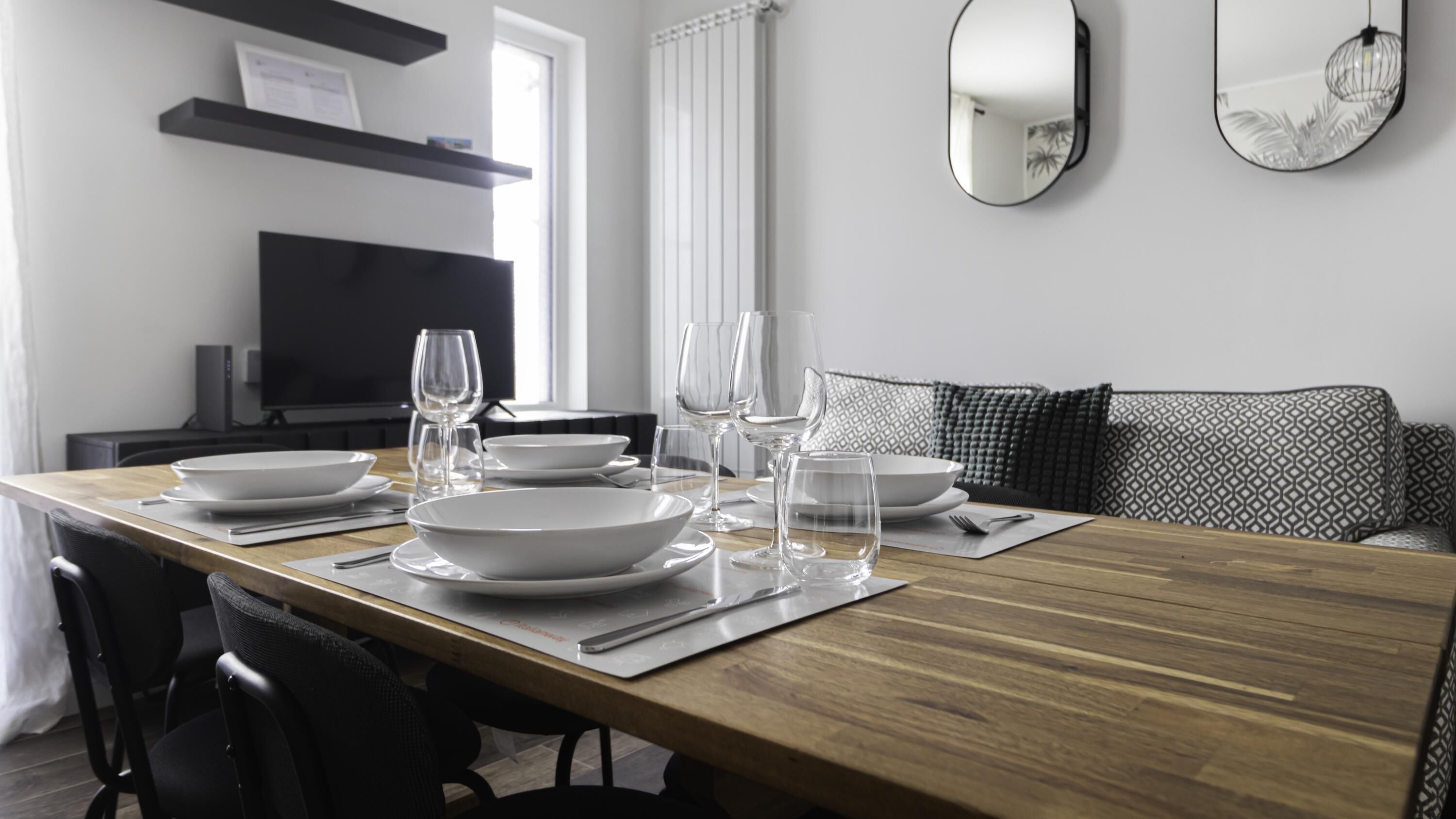 A dining table set with white plates, glasses, and cutlery, a black chair, and a patterned sofa in a modern room with a TV and shelves.