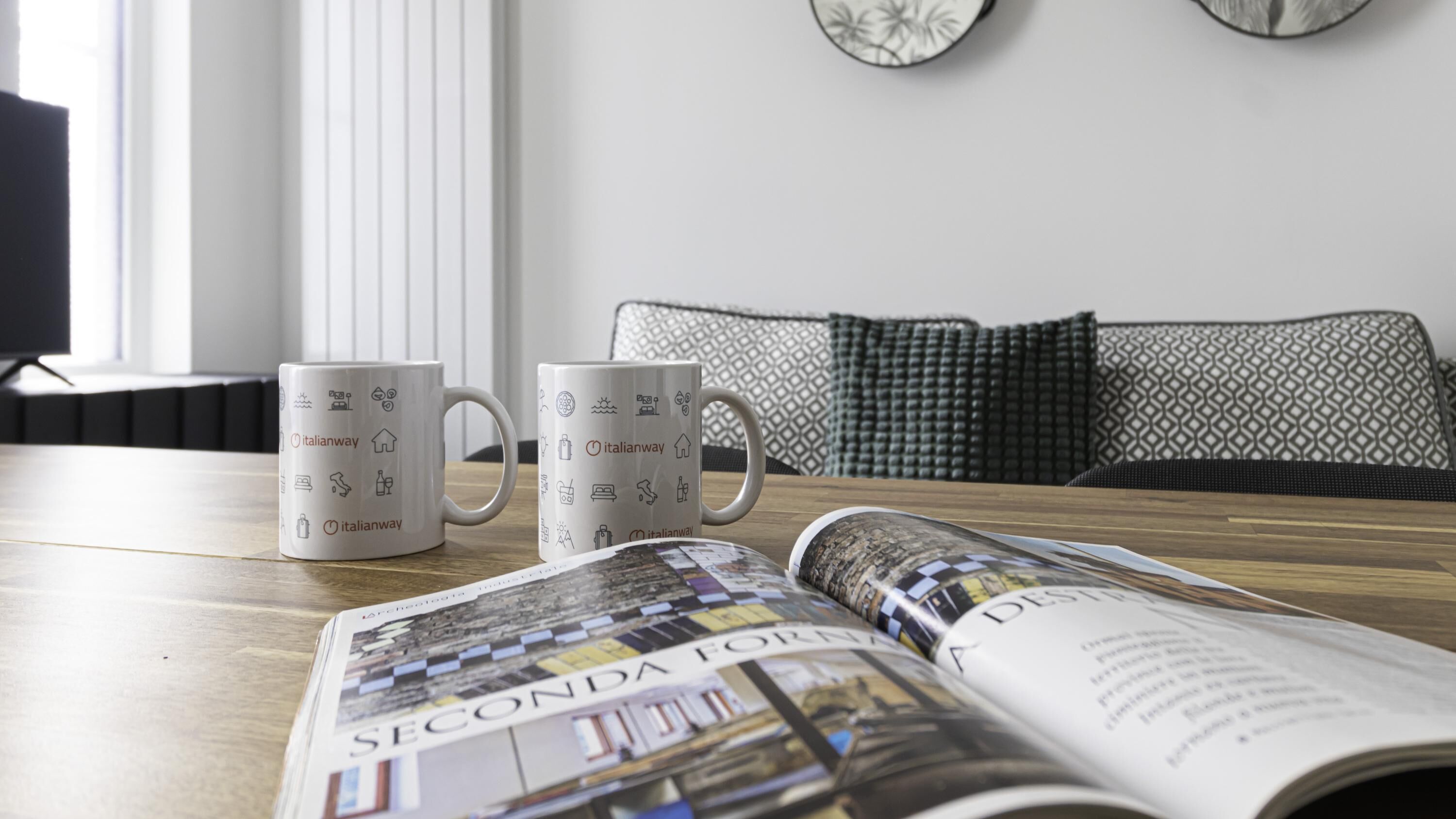 Two white mugs with Italianway logos on a wooden table, a magazine open to a page titled "Seconda Fornare," and a patterned sofa in the background.