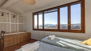 Une chambre avec deux lits, une commode en bois, une bibliothèque intégrée et une grande fenêtre donnant sur la ville.