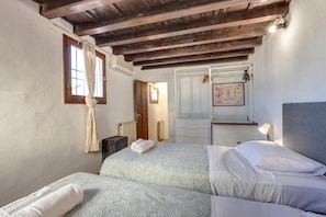 Een slaapkamer met twee bedden, houten balken in het plafond, een raam met gordijnen, een radiator en een deur met een afbeelding.
