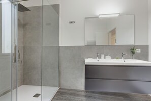 Une salle de bain moderne dotée d’une douche à parois de verre, d’un grand miroir et d’un meuble-lavabo blanc avec un plan de travail foncé.