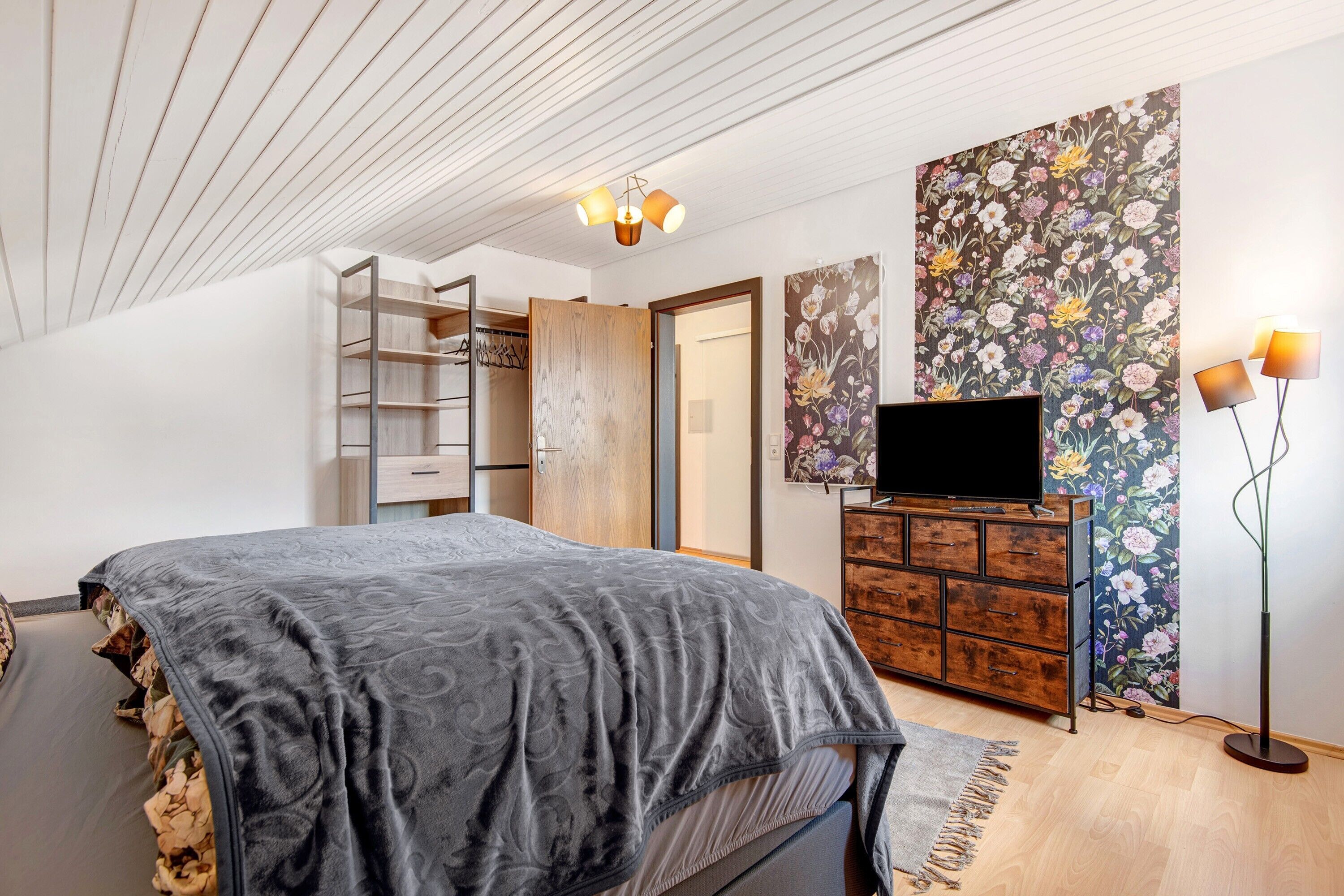 Un dormitorio con cama, televisor, armario, escritorio y lámpara de pie.