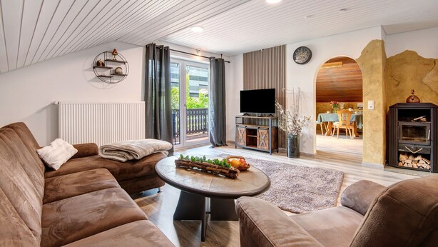 Een moderne woonkamer met een bruine bank, een ronde salontafel met een decoratief houtstuk, een open haard en een zichtbare eethoek door een boog.