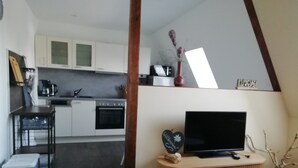 A modern kitchen with white cabinets, a stove, and a coffee maker. There is a TV on a wooden stand and a vase with flowers on the wall. - Apartment 'Am Froschteich' with Private Terrace, Shared Garden and Wi-Fi (Feldberger Seenlandschaft)