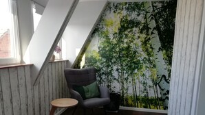A corner room with a sloped ceiling, a wooden bench, a small round table, a grey upholstered chair with a green cushion, and a wall mural of trees. - Apartment 'Am Froschteich' with Private Terrace, Shared Garden and Wi-Fi (Feldberger Seenlandschaft)