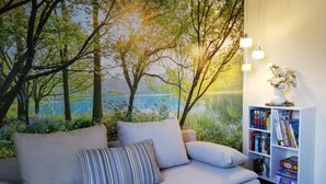 A living room with a sofa, pillows, a bookshelf, and a wall mural of a forest and lake. - Apartment 'Am Froschteich' with Private Terrace, Shared Garden and Wi-Fi (Feldberger Seenlandschaft)