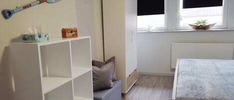 Une chambre avec un lit, une armoire blanche, une étagère avec une planche de paddle, un canapé gris et une fenêtre avec des stores.