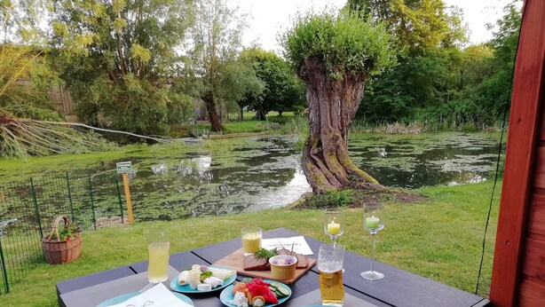 An outdoor dining table with plates, cutlery, and drinks, set against a backdrop of a pond and trees. - Apartment 'Am Froschteich' with Private Terrace, Shared Garden and Wi-Fi (Feldberger Seenlandschaft)