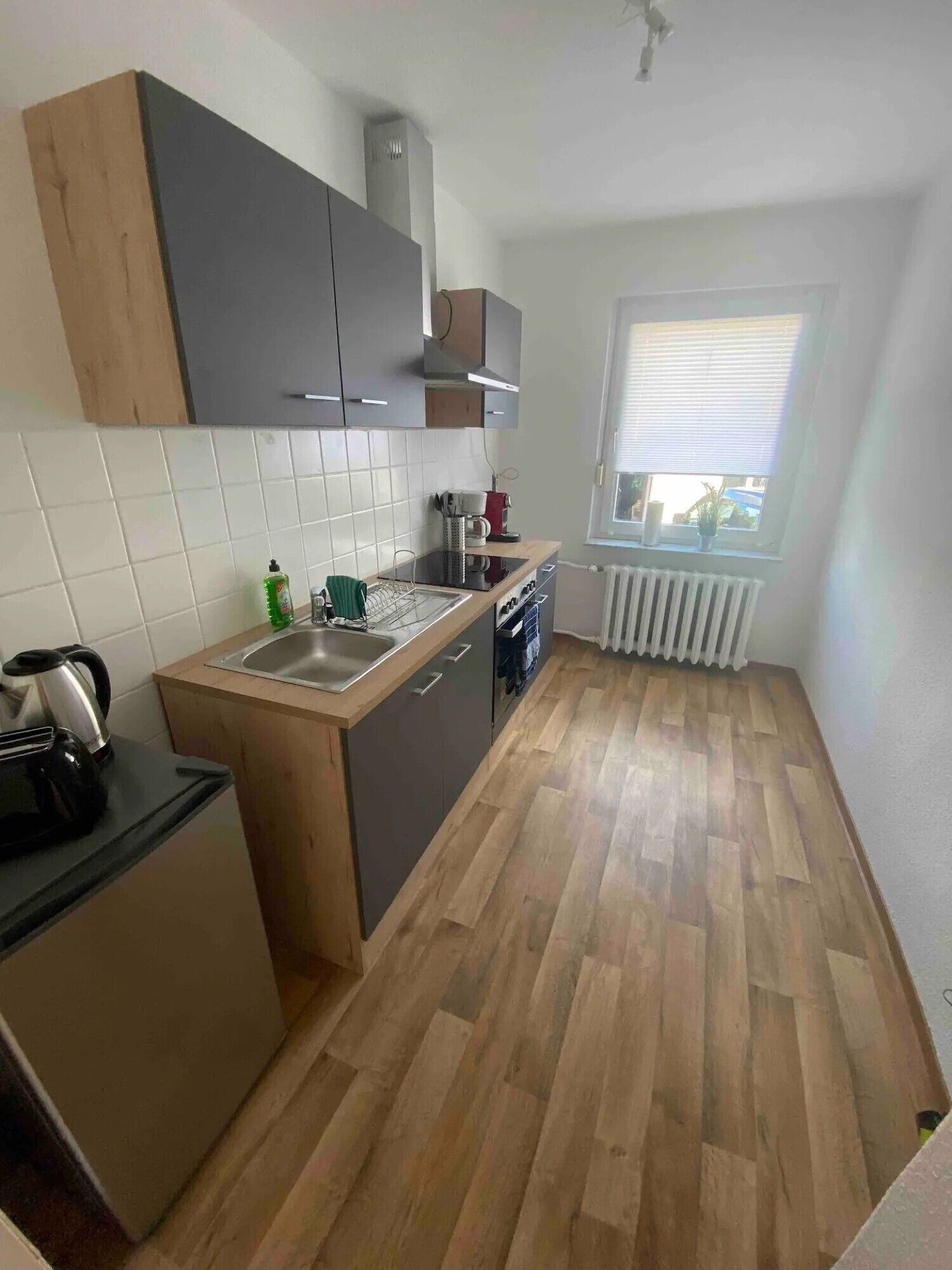 Una cocina compacta con un lavamanos, estufa y armarios, una ventana con persianas y un radiador.