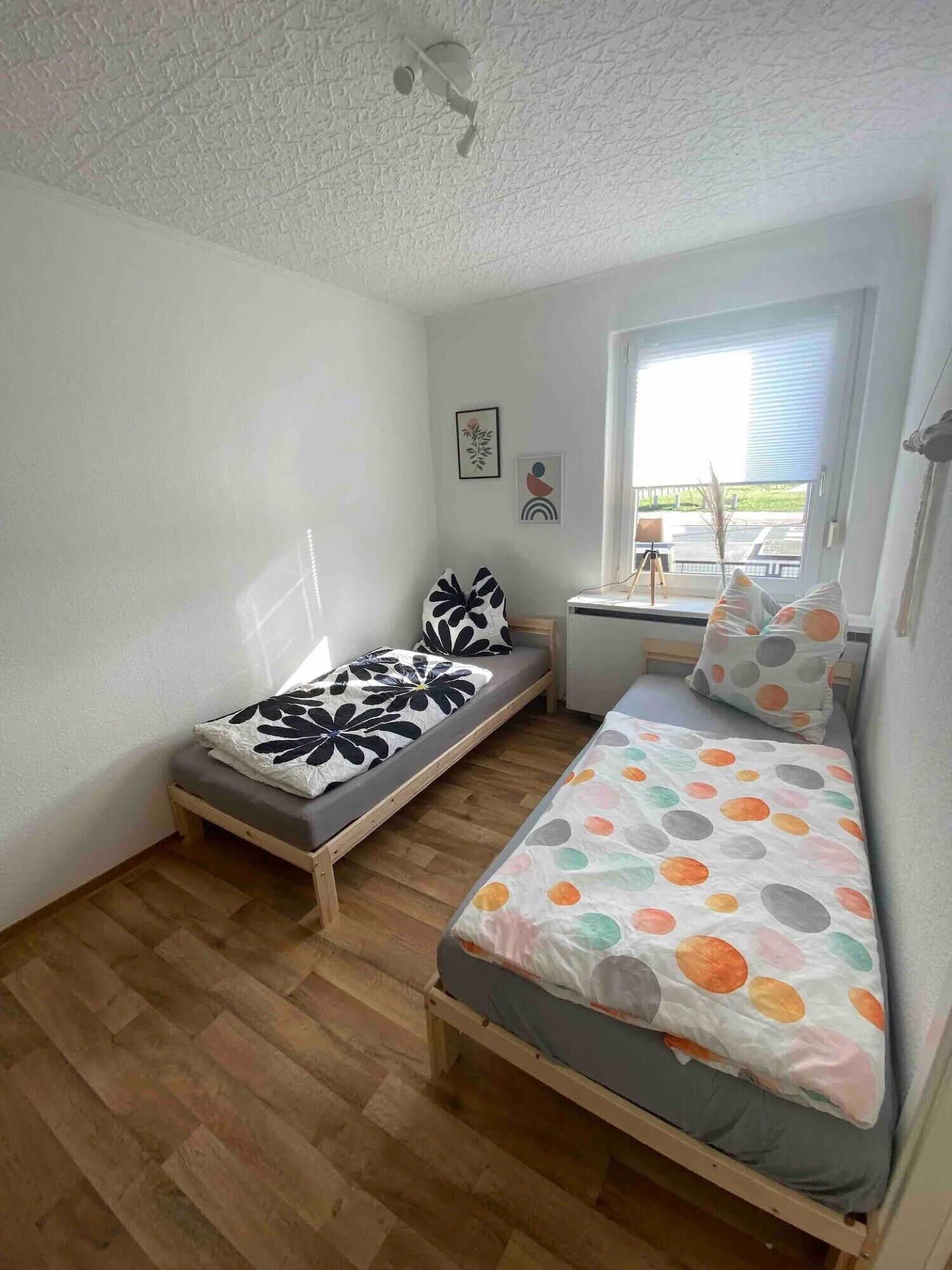 A small, well-lit room with two single beds, a window with blinds, and a ceiling fan.