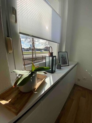 Una ventana con vista a una carretera y una cerca, una planta en maceta, un cuadro enmarcado y un objeto metálico sobre una encimera.