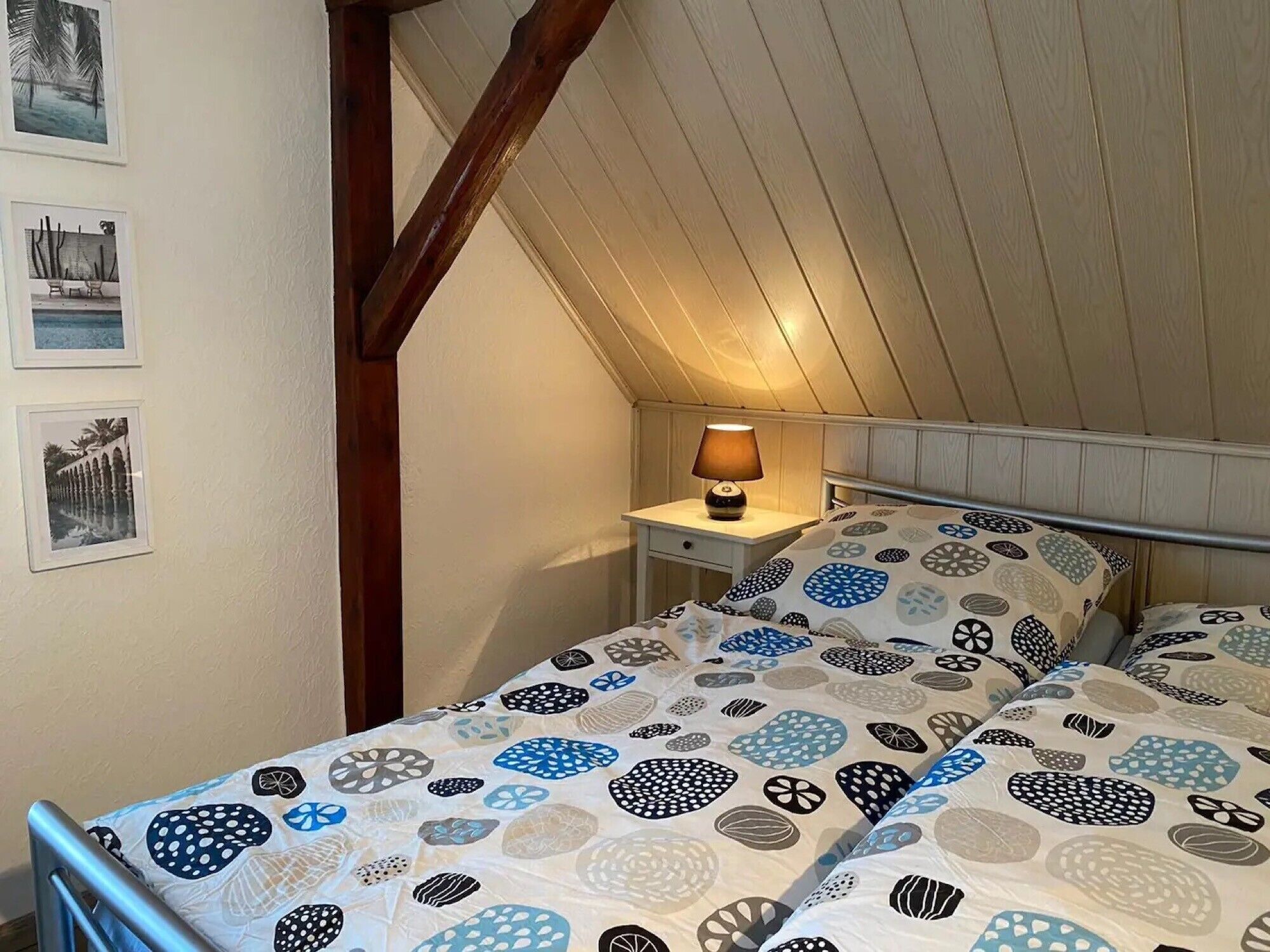Un dormitorio con techo inclinado, una cama con ropa de cama estampada, una mesita de noche con lámpara y cuadros en la pared.