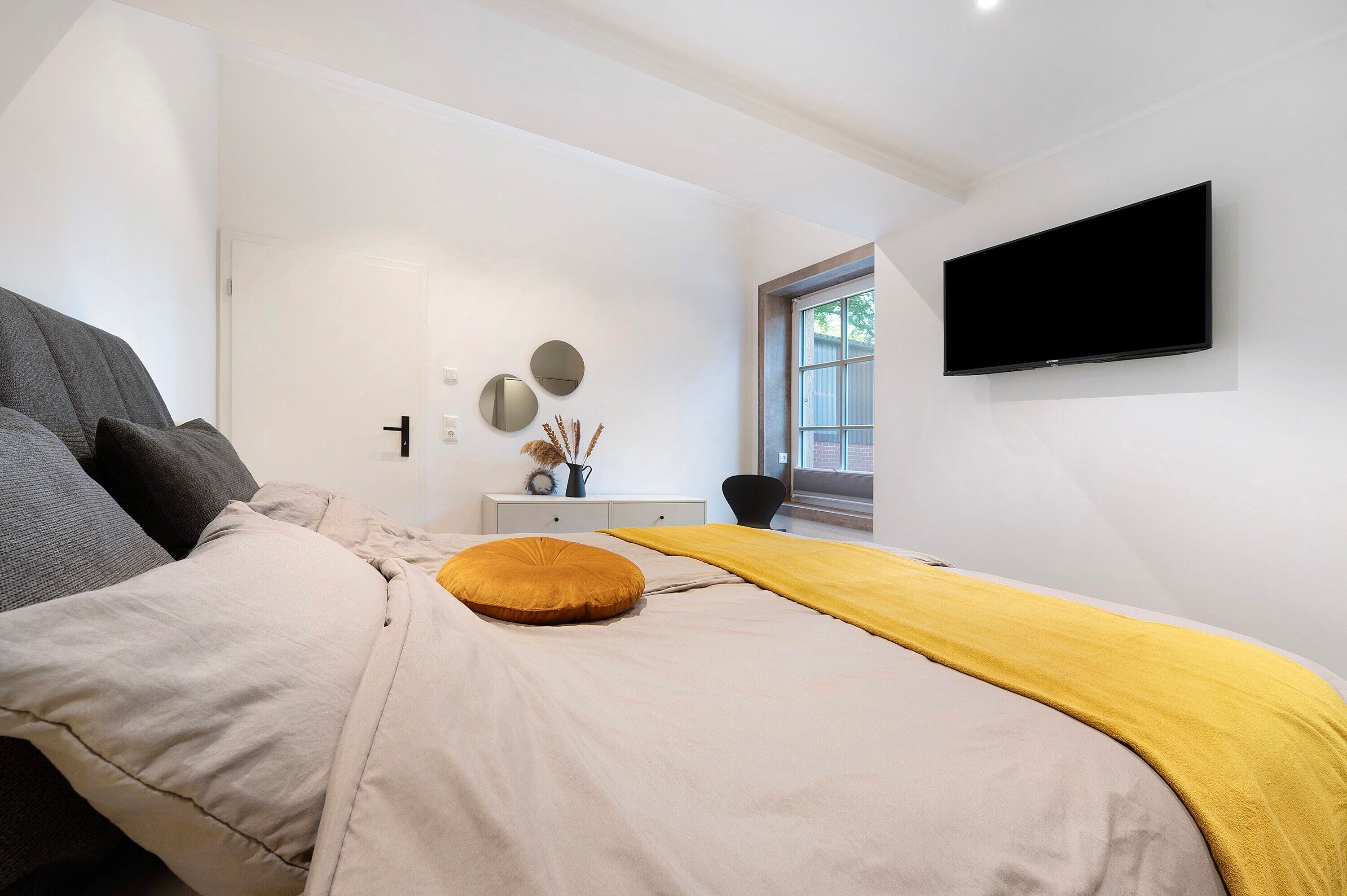 Ein modernes Schlafzimmer mit Bett, Flachbildfernseher, Fenster und kleinem Schreibtisch mit Stuhl.