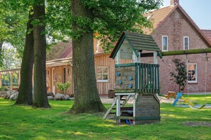 A wooden playhouse with climbing features, a swing set, and a brick building in the background.