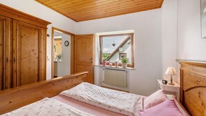 Une chambre avec un plafond en bois, une fenêtre donnant sur un immeuble, un lit avec une literie rose et une table de chevet avec une lampe.