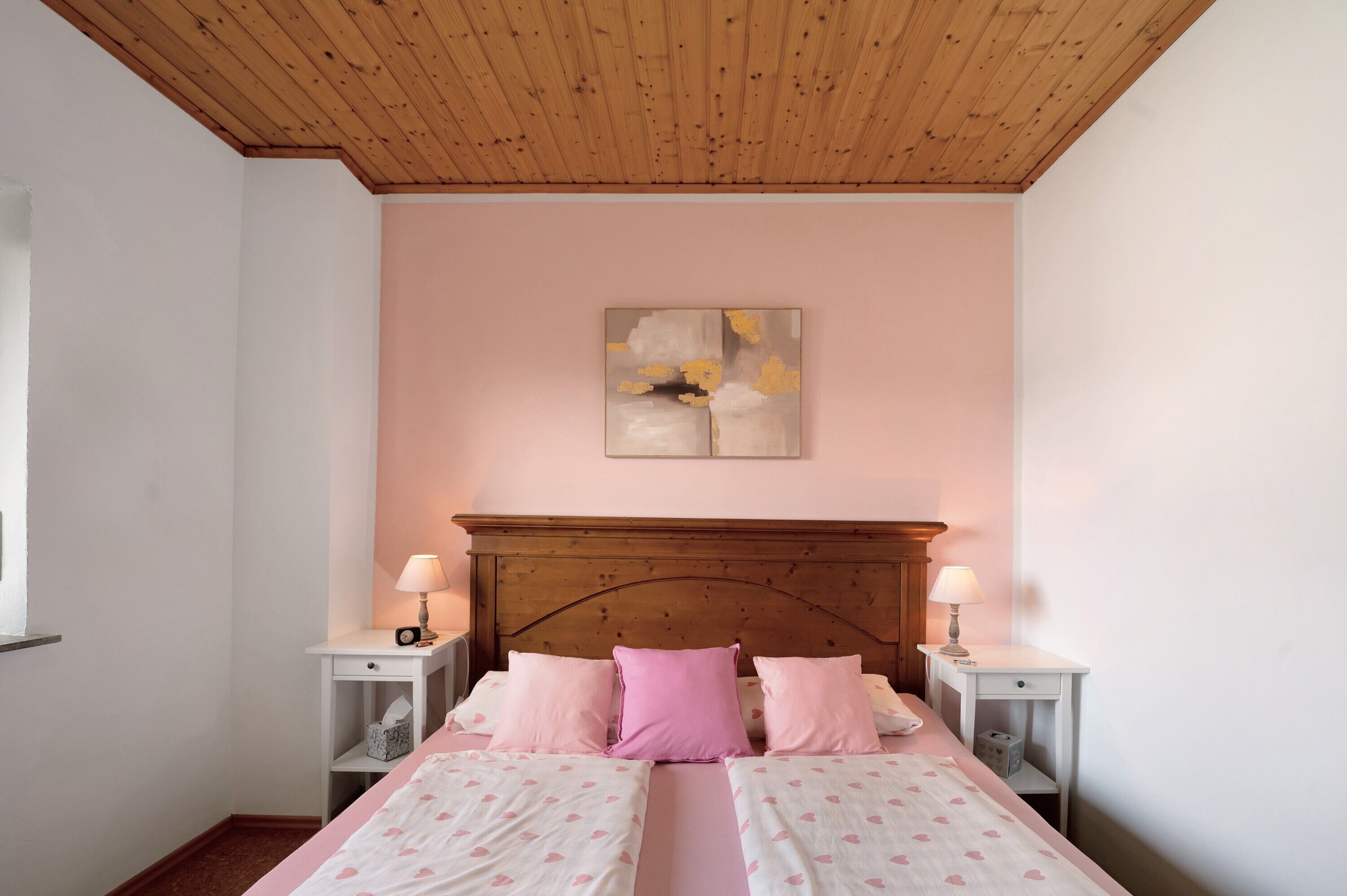 Ein Schlafzimmer mit Holzbalkendecke, einem Bett mit rosa Bettwäsche, zwei Nachttischen mit Lampen und einem an der Wand hängenden abstrakten Gemälde.