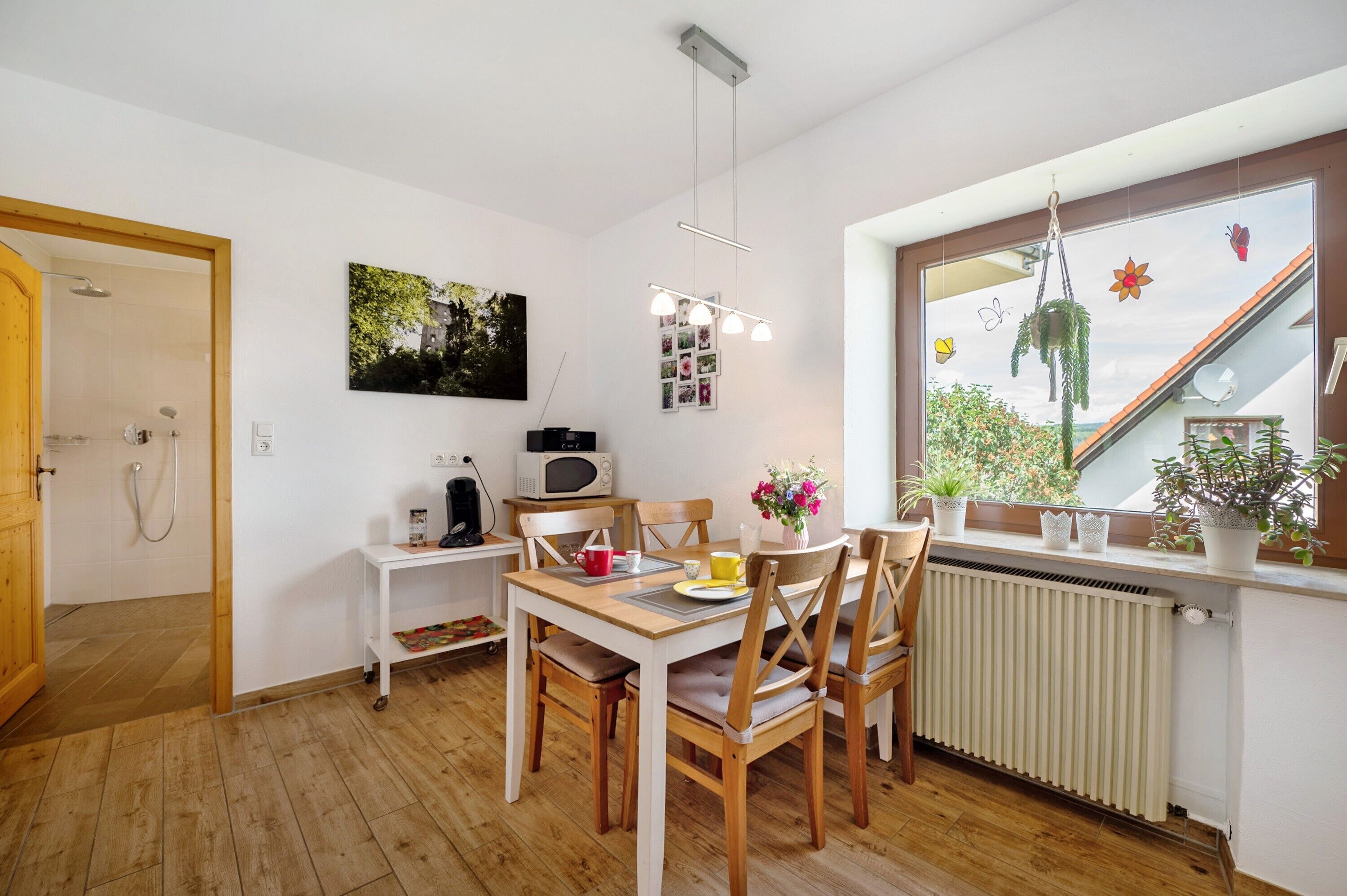 Cocina compacta con comedor, microondas, una mesita, sillas, radiador y ventana con plantas.