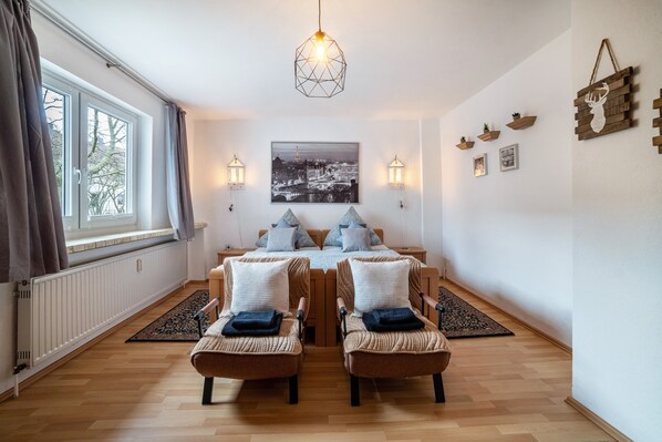 A room with a wooden floor, two armchairs, a table with chairs, a hanging light fixture, a framed cityscape picture, wall-mounted sconces, and a radiator.