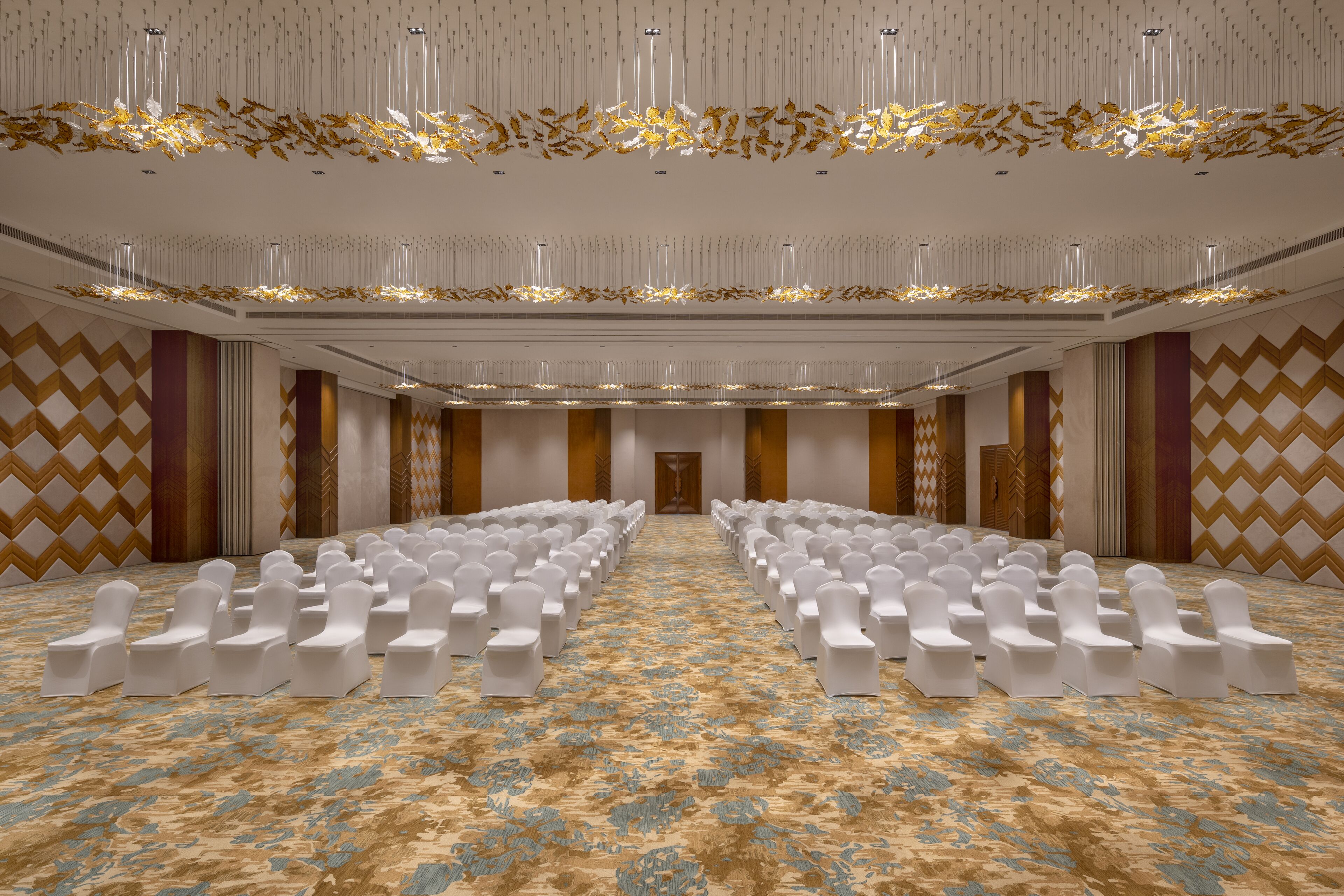 Een ruime congreszaal met rijen witte stoelen, een kamerbreed tapijt met dessin en decoratieve elementen aan het plafond.