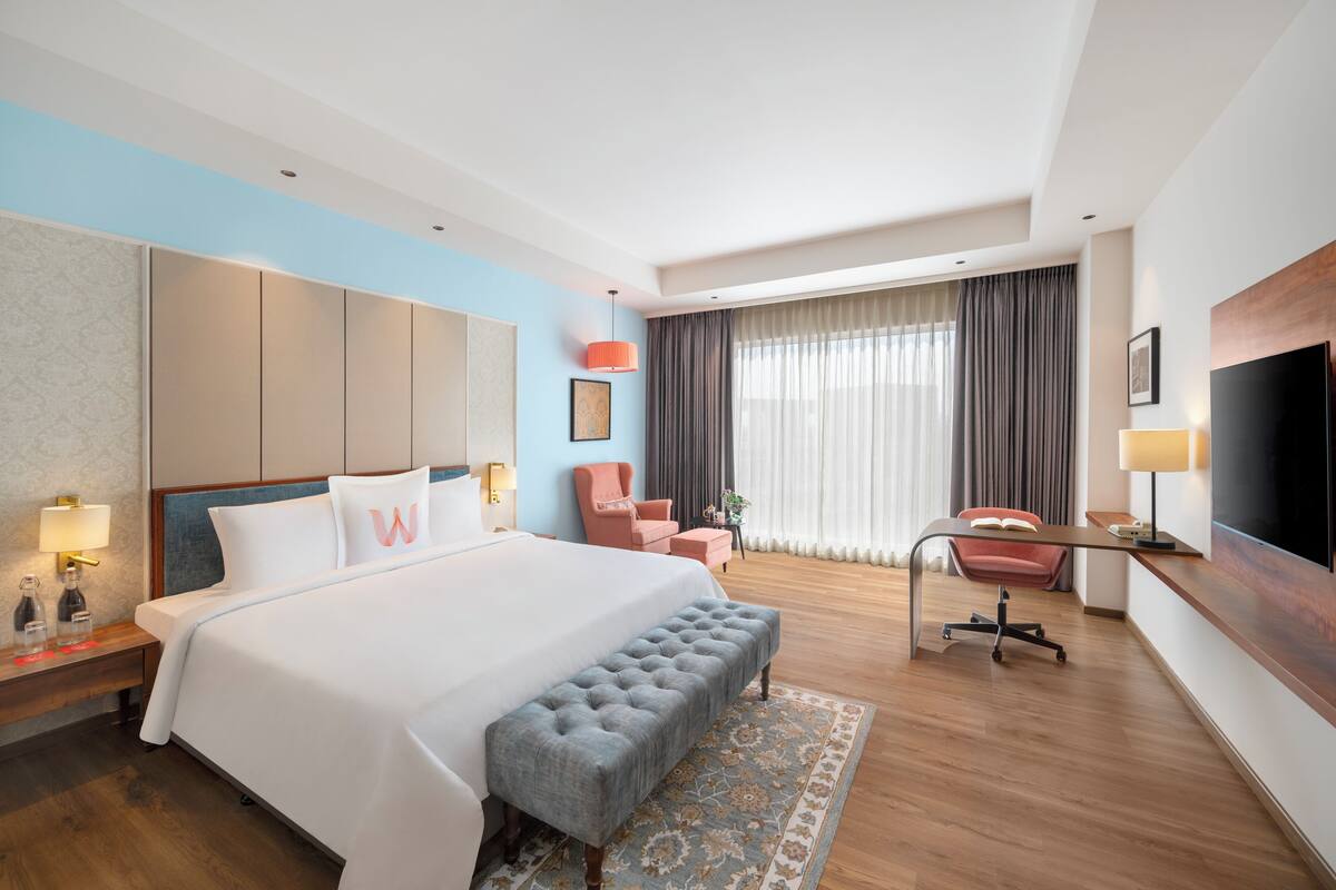Habitación de hotel con una cama grande, un escritorio, un televisor y una zona de estar con una silla y un reposapiés.