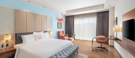 Habitación de hotel con una cama grande, un escritorio, un televisor y una zona de estar con una silla y un reposapiés.