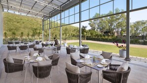 An outdoor dining area with wicker chairs and tables, large windows, and a view of a baseball field. - Welcomhotel By ITC Hotels Belagavi (Belagavi)